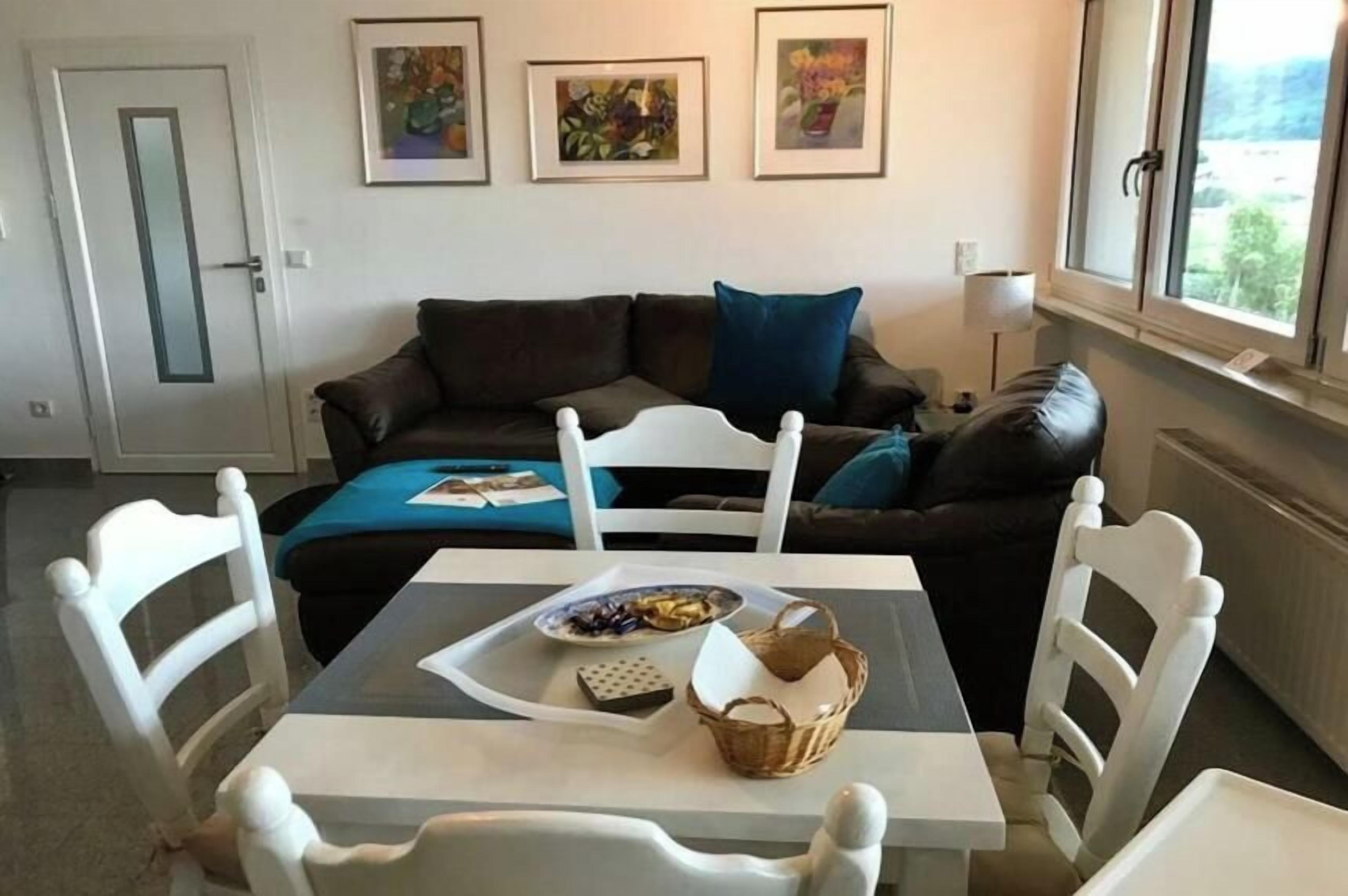Un salon avec un canapé marron, des chaises blanches, une table sur laquelle se trouvent un panier et une assiette de nourriture, et des tableaux encadrés accrochés au mur.