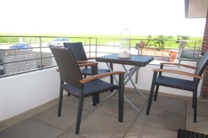 A balcony with two chairs and a table, a lantern, and a view of a parking area and greenery. - Residence Naturdüne Holiday Apartment No. 5 (Cuxhaven Duhnen)