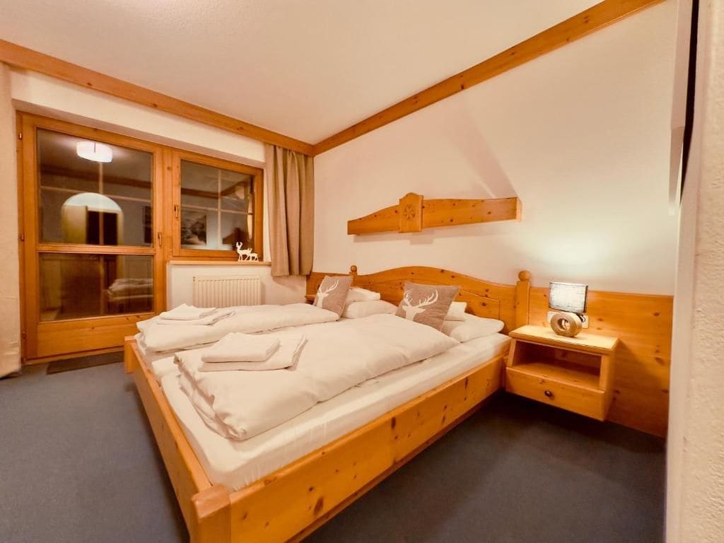 Een houten bed met hoofdeinde, een nachtkastje met een lamp en een raam met gordijnen.