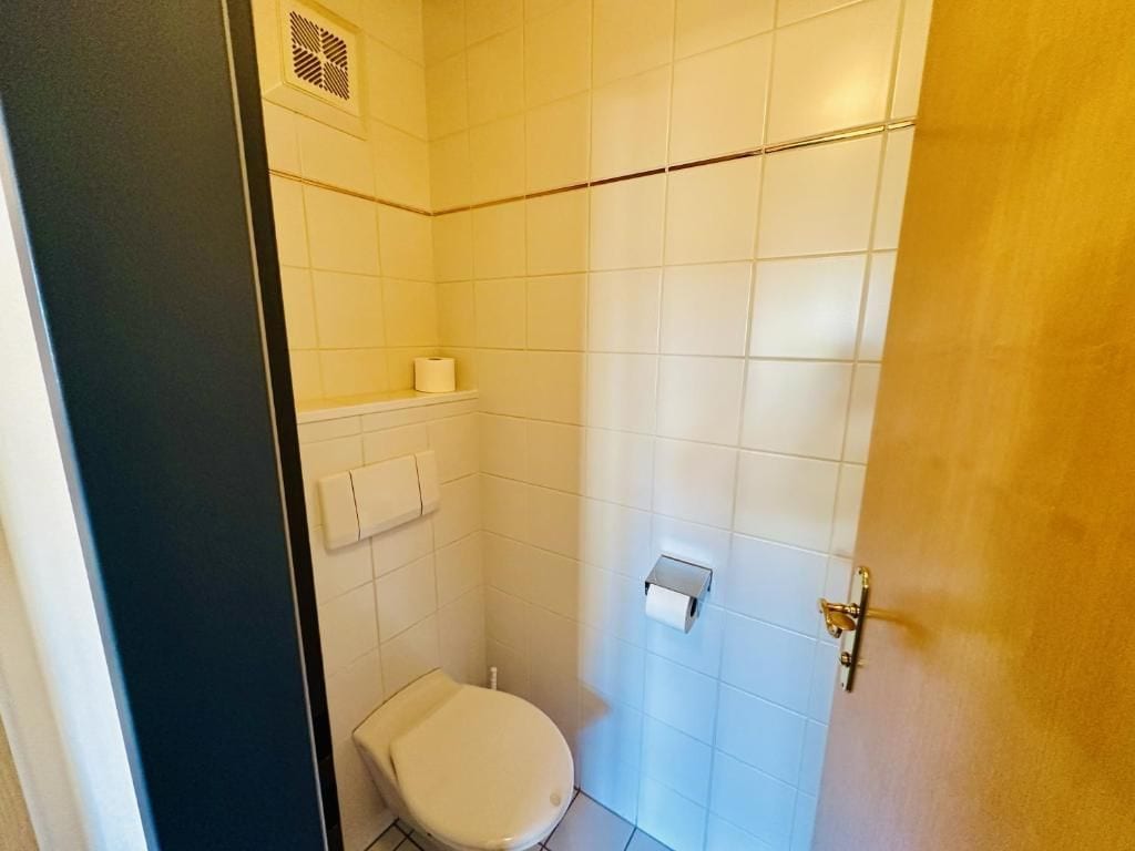 A small, compact bathroom with white tiles, a toilet, a towel rack, and a toilet paper holder.