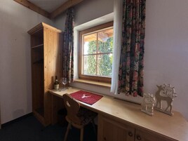 Una habitación con un escritorio de madera, una silla, un ventanal con cortinas y un estante con adornos.