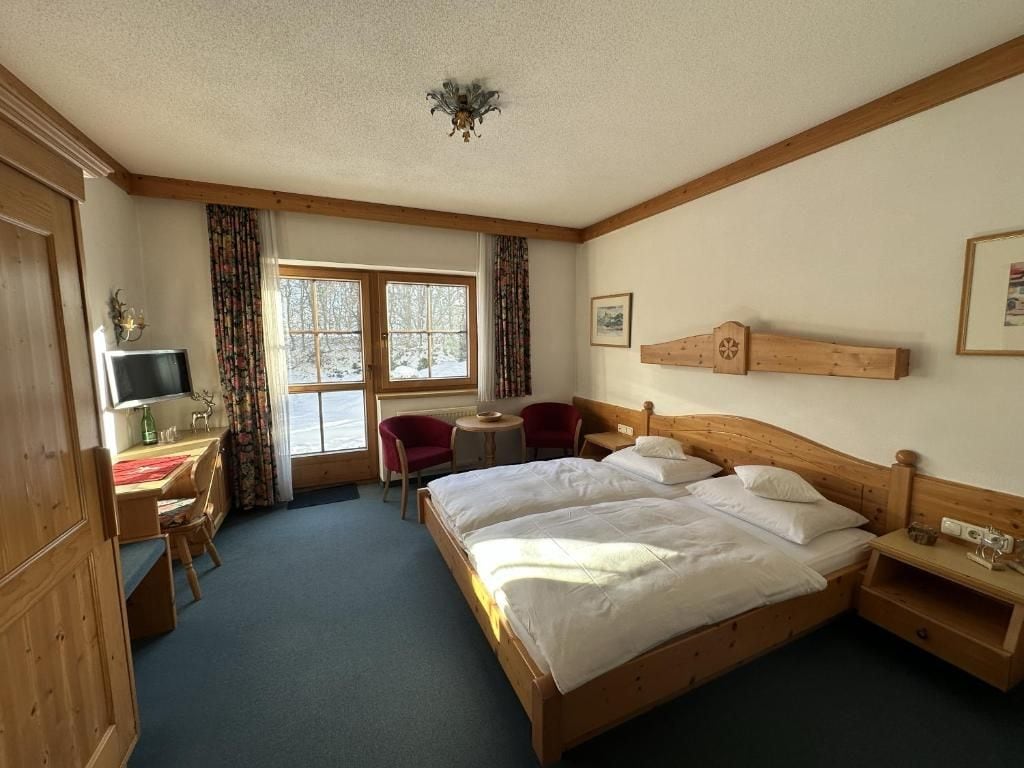 Une chambre d’hôtel avec un lit en bois, un bureau avec une chaise, une petite table et une fenêtre avec des rideaux.