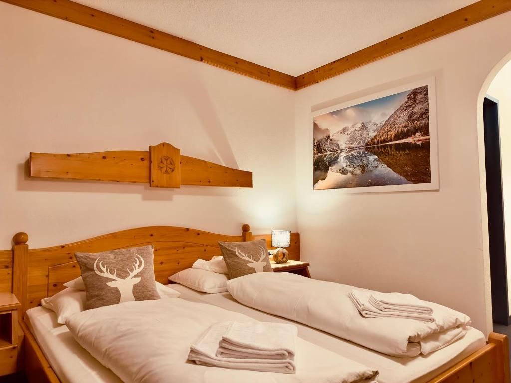 Un lit double avec du linge de lit blanc, deux oreillers ornés de motifs de cerfs, une tête de lit en bois, une table de chevet avec une lampe et un tableau représentant un paysage montagneux.