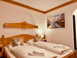 Una cama doble con sábanas blancas, dos almohadas con diseño de ciervo, un cabecero de madera, una mesita de noche con lámpara y un cuadro con paisaje montañoso.