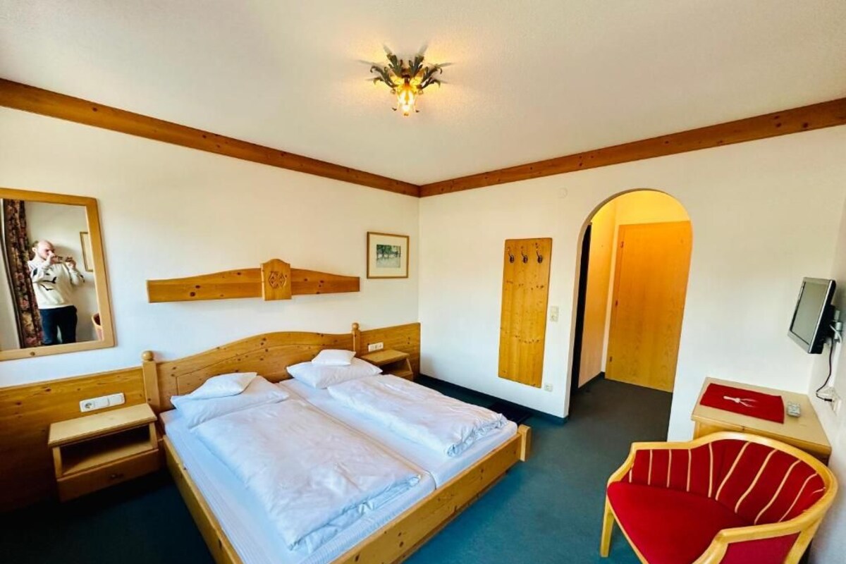 Ein Zimmer mit einem hölzernen Bett, einem roten Sessel, einem Schreibtisch aus Holz, einem Spiegel und einer Tür.
