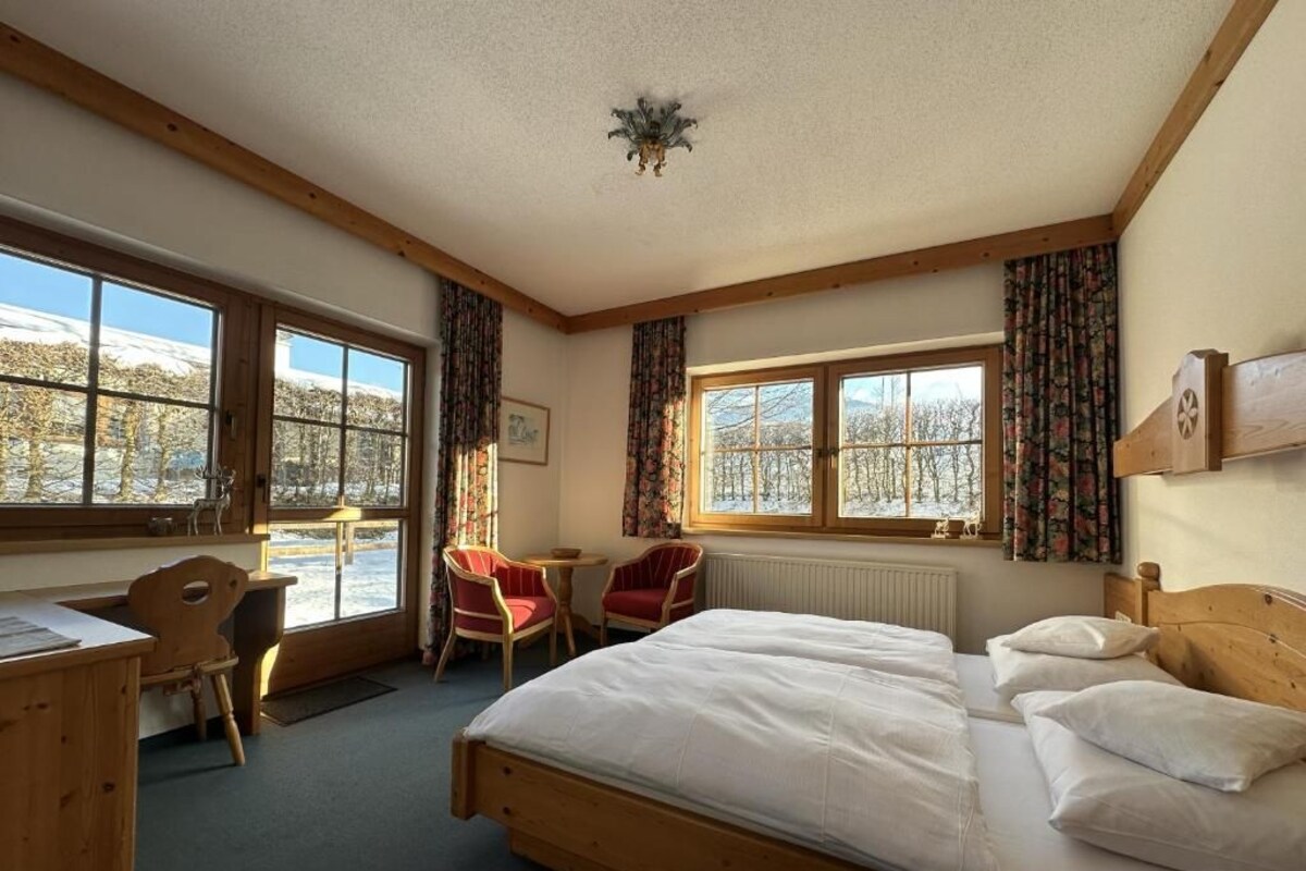 Ein Zimmer mit einem Bett, einem Schreibtisch, einem Stuhl und zwei fensterförmigen Öffnungen mit Vorhängen.
