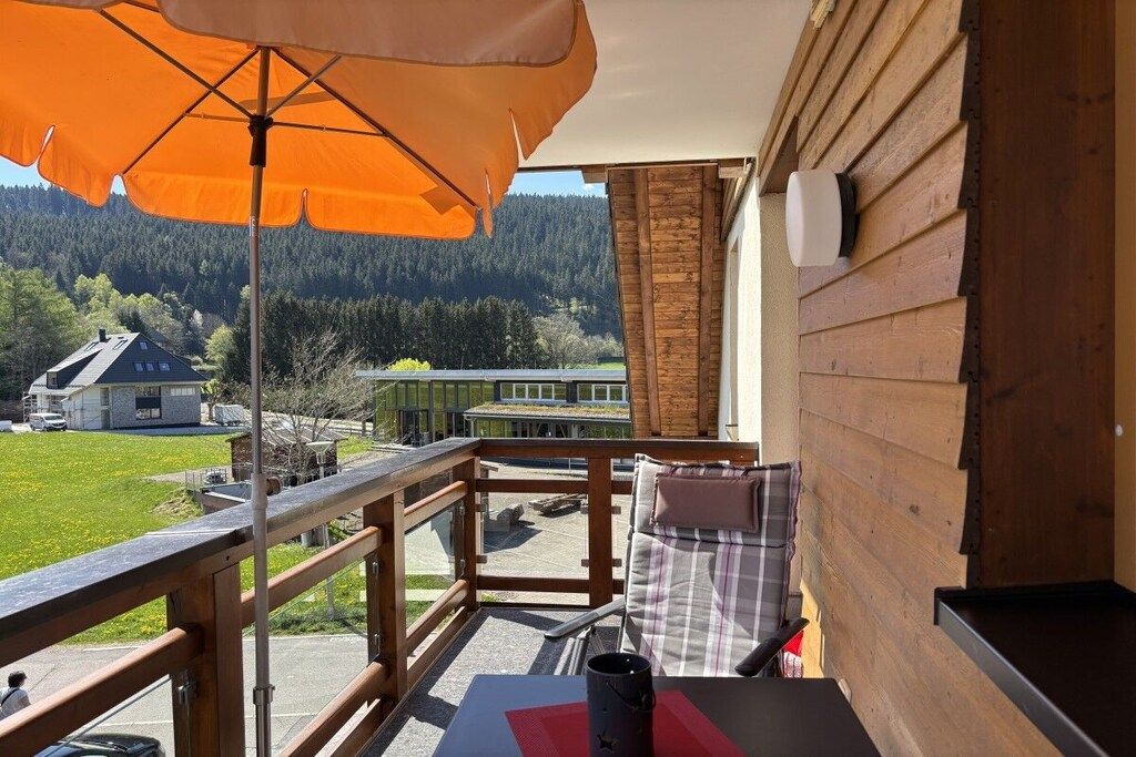 Een balkon met een houten leuning, een tafel met een zwart voorwerp, en uitzicht op een groen huis en bos.