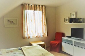 Ein Zimmer mit einem Bett, einem kleinen Tisch, einem roten Sessel, einem Fernseher auf einer Kommode und einem Fenster mit Vorhängen.