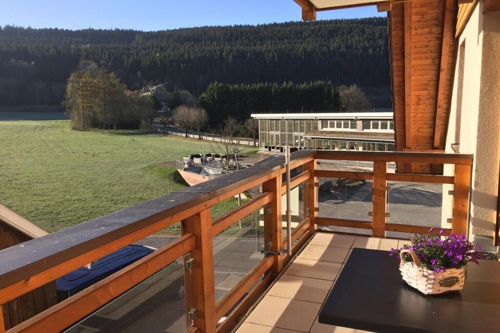 Ein Balkon mit Holzgeländer, der auf ein Wiesenfeld und ein Gebäude in der Ferne blickt.