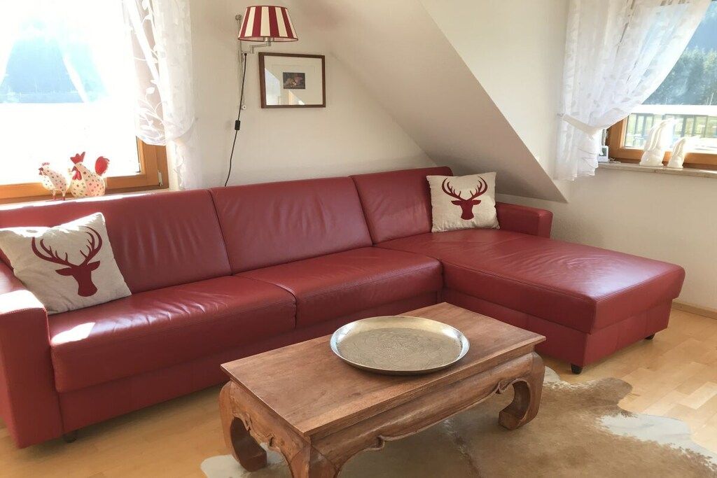 Een rode leren bank met een houten salontafel in een kamer met een schuine kap en een raam met gordijnen.