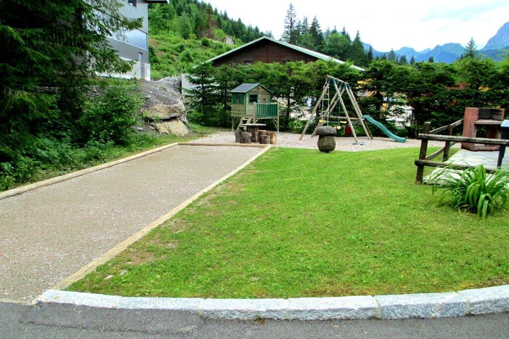 A playground with a swing set and slide, surrounded by a grassy area and a building with a sloped roof.