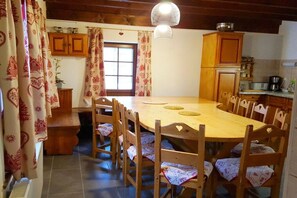 Un coin repas avec une table et des chaises en bois, une fenêtre avec des rideaux et un espace cuisine équipé de placards et d’appareils électroménagers.