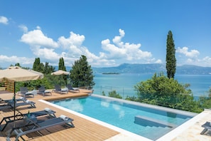 A poolside area with lounge chairs, a wooden deck, and a view of the sea and mountains. - Villa Nera Corfu (Kerkira)