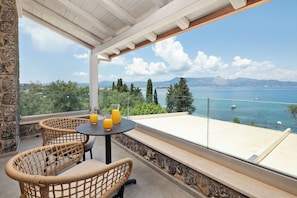A balcony with wicker chairs and a table, overlooking a sea with boats and mountains in the distance. - Villa Nera Corfu (Kerkira)