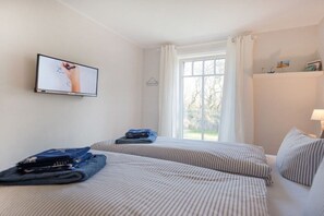 Ein Hotelzimmer mit zwei Betten, einem an der Wand befestigten Fernseher, einem Fenster mit Vorhängen und einem Regal mit Dekoration.