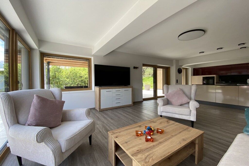Ein großzügiger Wohnbereich mit Couchtisch aus Holz, zwei Polsterstühlen, einem Flachbildfernseher und einer Küche im Hintergrund.