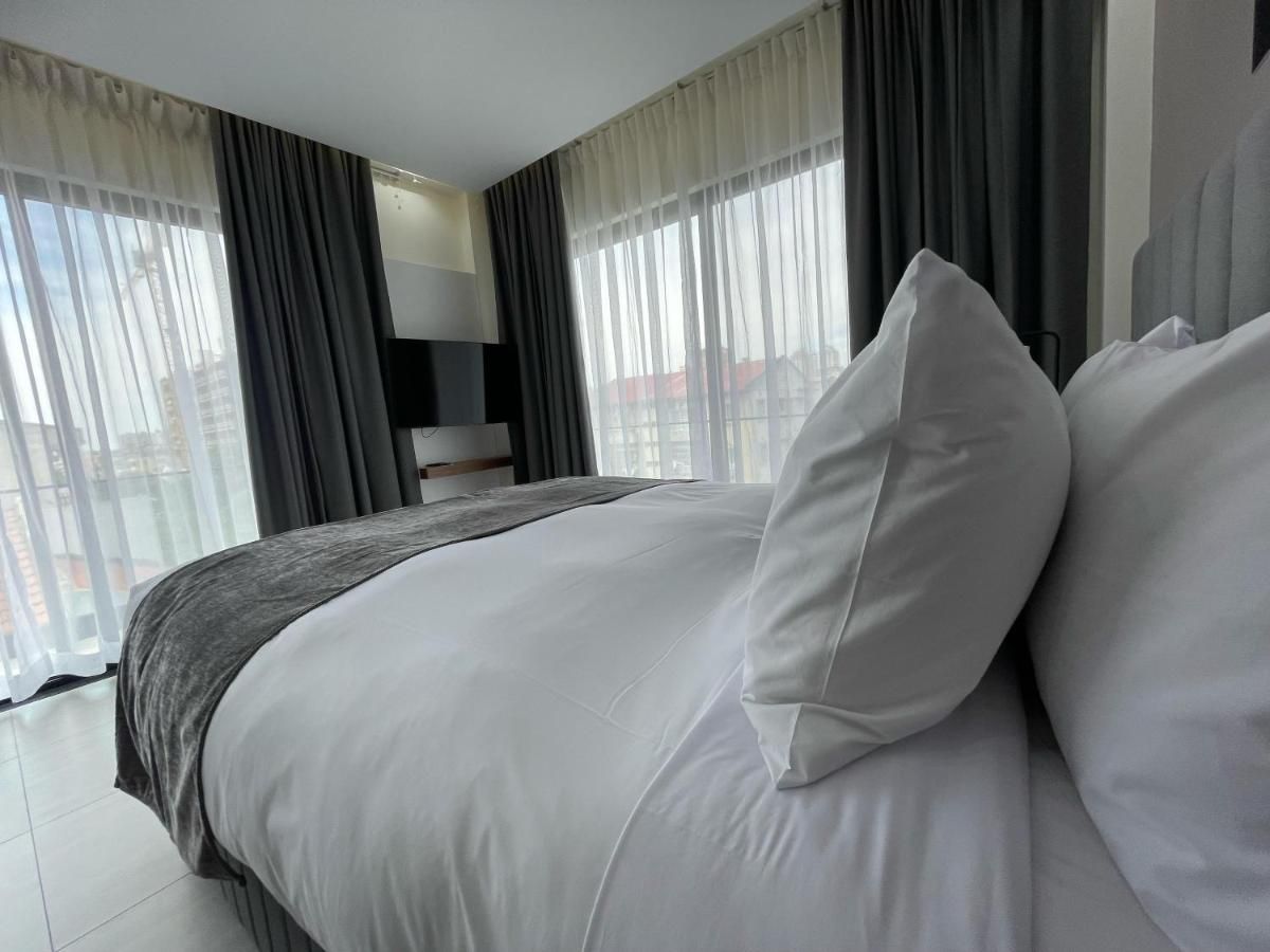 A hotel room with a large bed, a flat-screen TV, and a view of the city through sheer curtains.