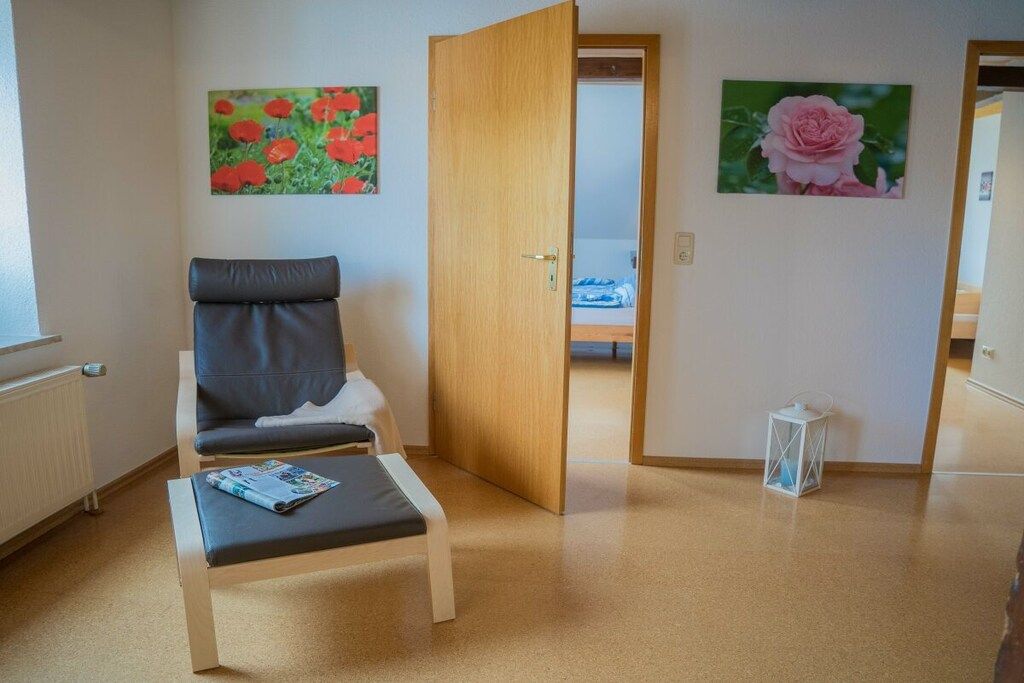 Een kamer met een stoel, een klein tafeltje en een deur naar een andere ruimte. Op de muren hangen twee schilderijen.