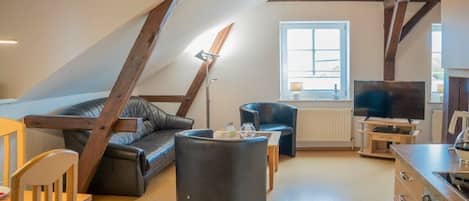 A cozy living room with a sloped ceiling, a black leather sofa, a round ottoman, a television, and a dining area with a table and chairs.