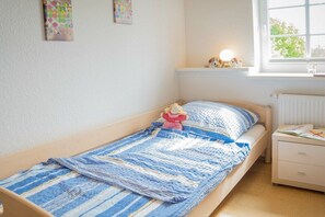 A single bed with a striped blanket, a nightstand with a lamp, and a window with a view of greenery. - Holiday apartment in Fehmarn with Wi-Fi (Fehmarn)