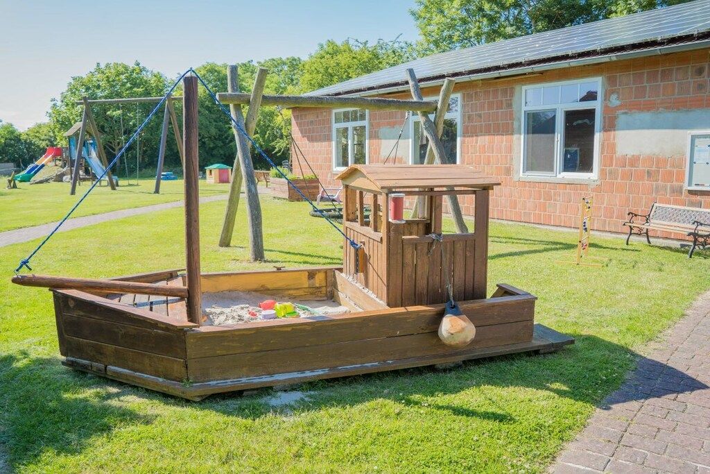 A wooden play structure in a grassy area with a brick building in the background.