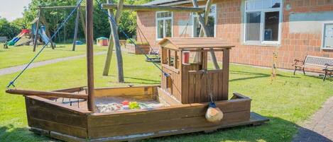 A wooden play structure in a grassy area with a brick building in the background.