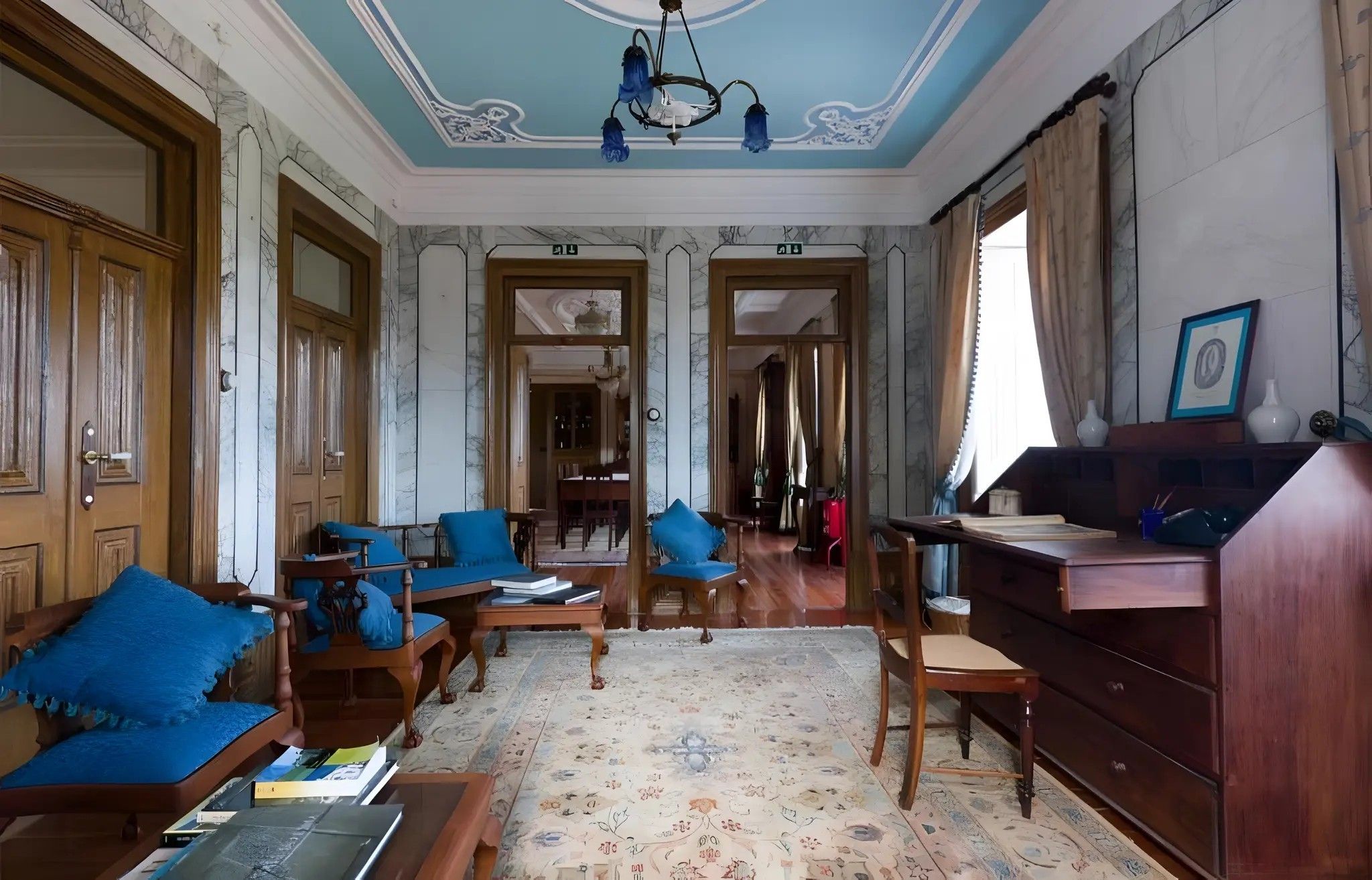 A room with a wooden desk, blue upholstered chairs, a patterned carpet, and a chandelier.