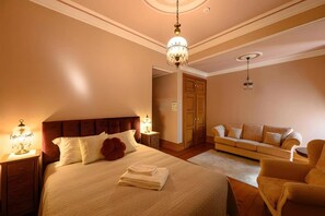 A hotel bedroom with a large bed, bedside tables, a sofa, and a chandelier.