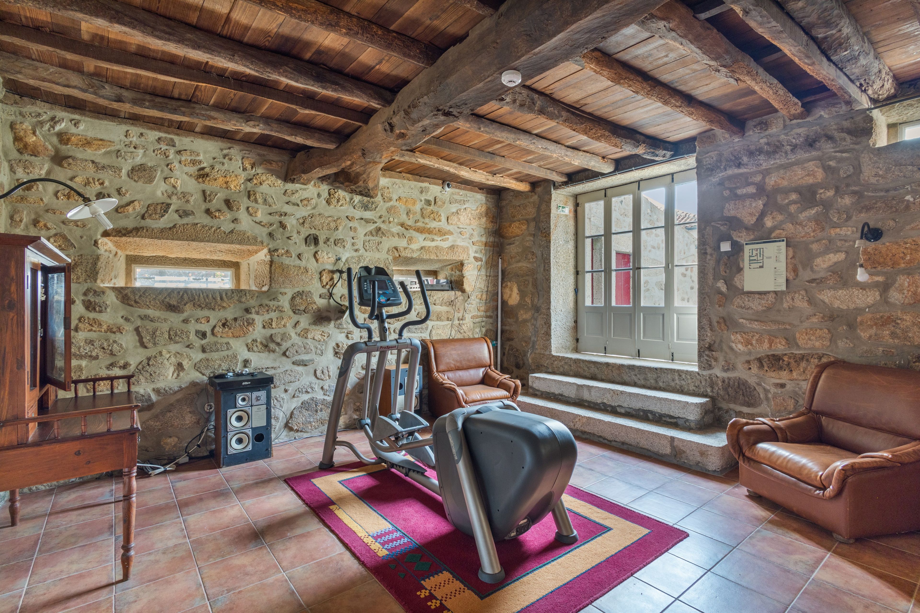 A rustic room with stone walls, a wooden ceiling, a treadmill, a leather sofa, and a window with a view.