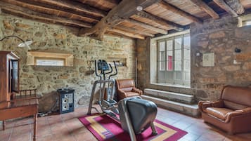 A rustic room with stone walls, a wooden ceiling, a treadmill, a leather sofa, and a window with a view.