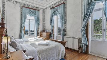 A bedroom with two beds, a wooden dresser, a nightstand, a lamp, and a view of the outdoors through large windows.