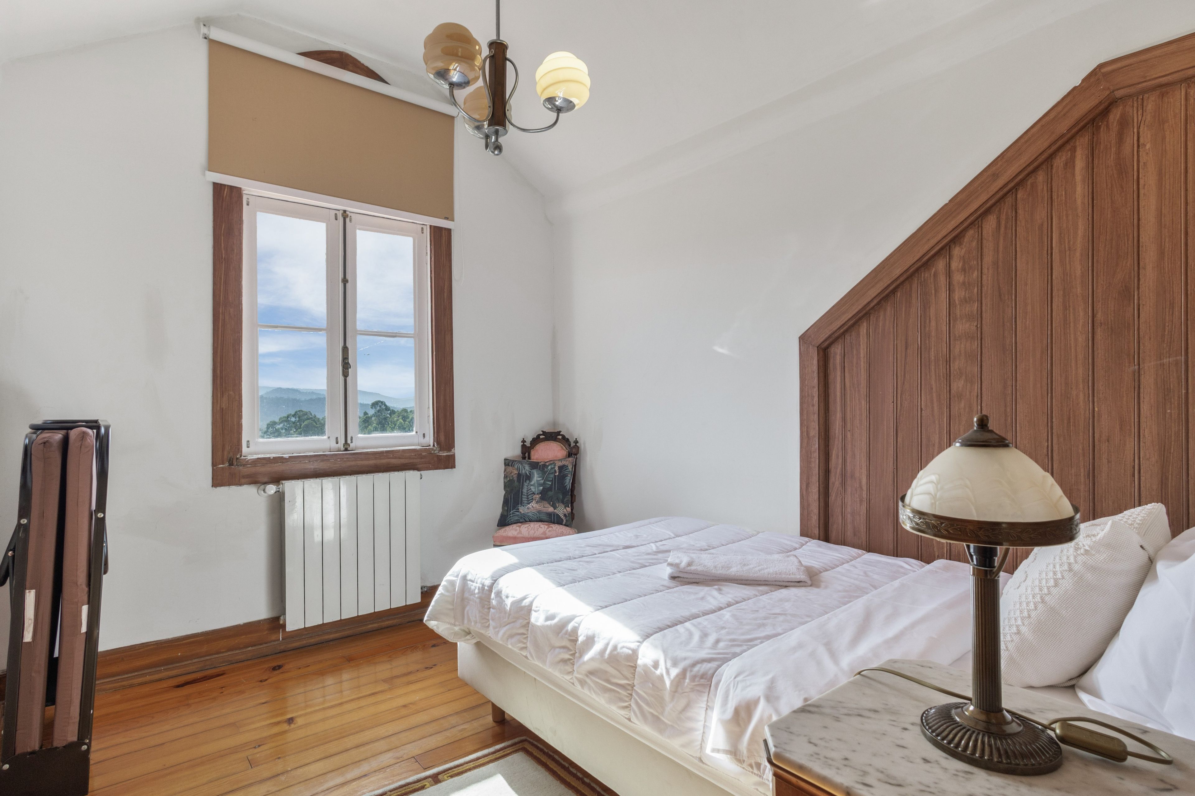 Une chambre avec un plancher en bois, un lit avec une literie blanche, une table de chevet, une lampe, une fenêtre donnant sur un paysage agréable et un radiateur.