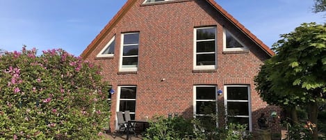 Una casa de ladrillo con techo triangular, un porche delantero y un jardín con flores rosadas.