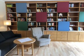 A library with bookshelves, a grey armchair, a round wooden table, and a dark sofa. - Bründermann holiday apartment (Edewecht)