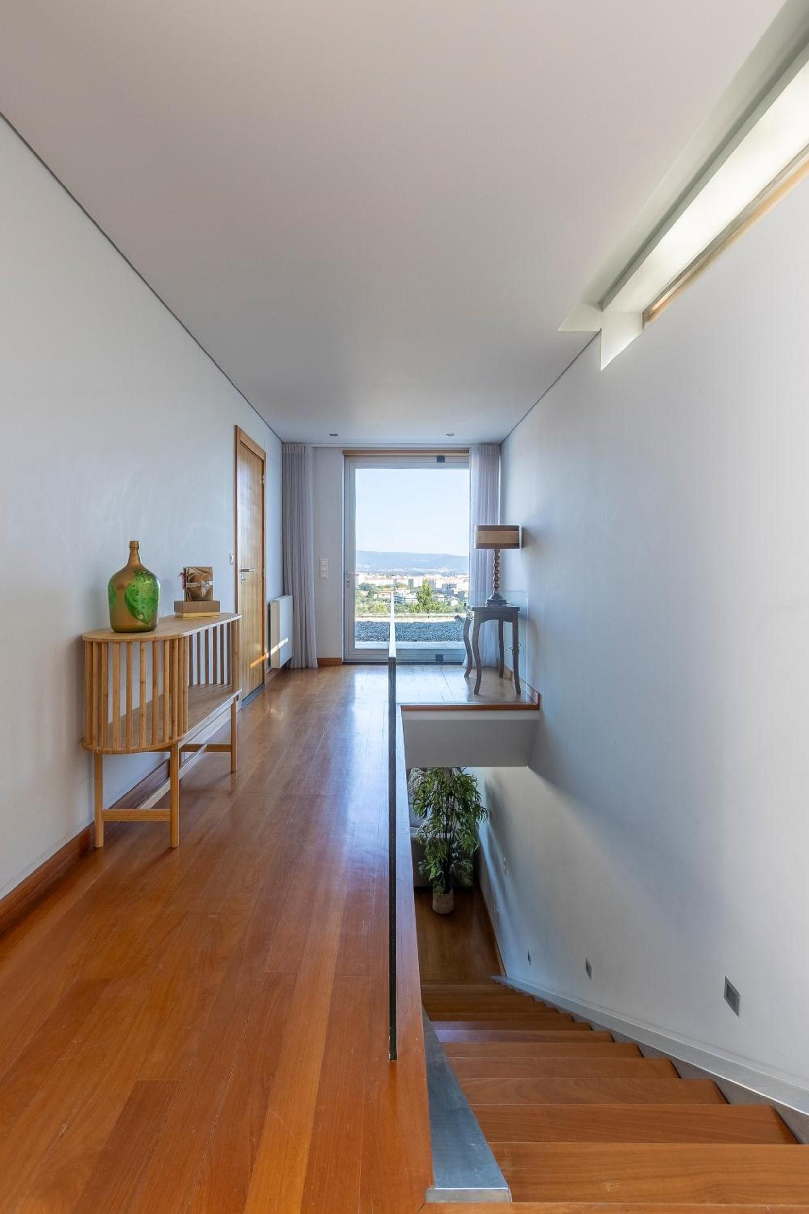 Un interior moderno con suelo de madera, una escalera, vistas a través de un vidrio y una pequeña mesa con lámpara.