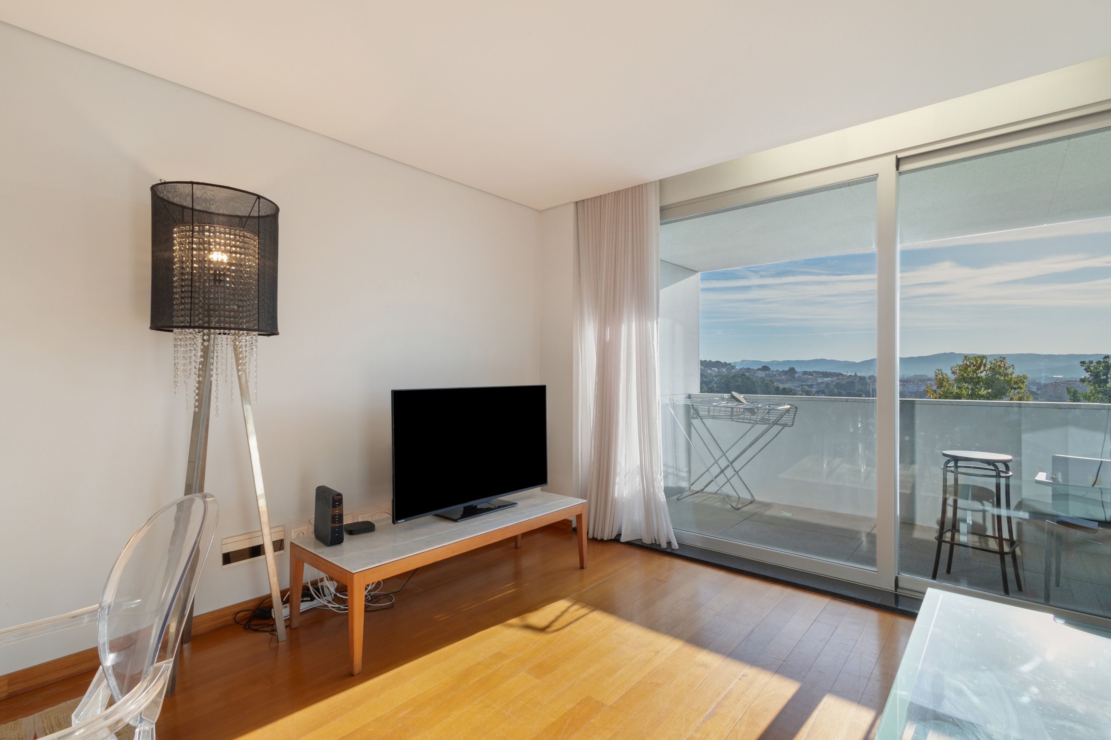Un soggiorno moderno con una TV a schermo piatto su un supporto in legno, una lampada da terra e un tavolino in vetro con una sedia.