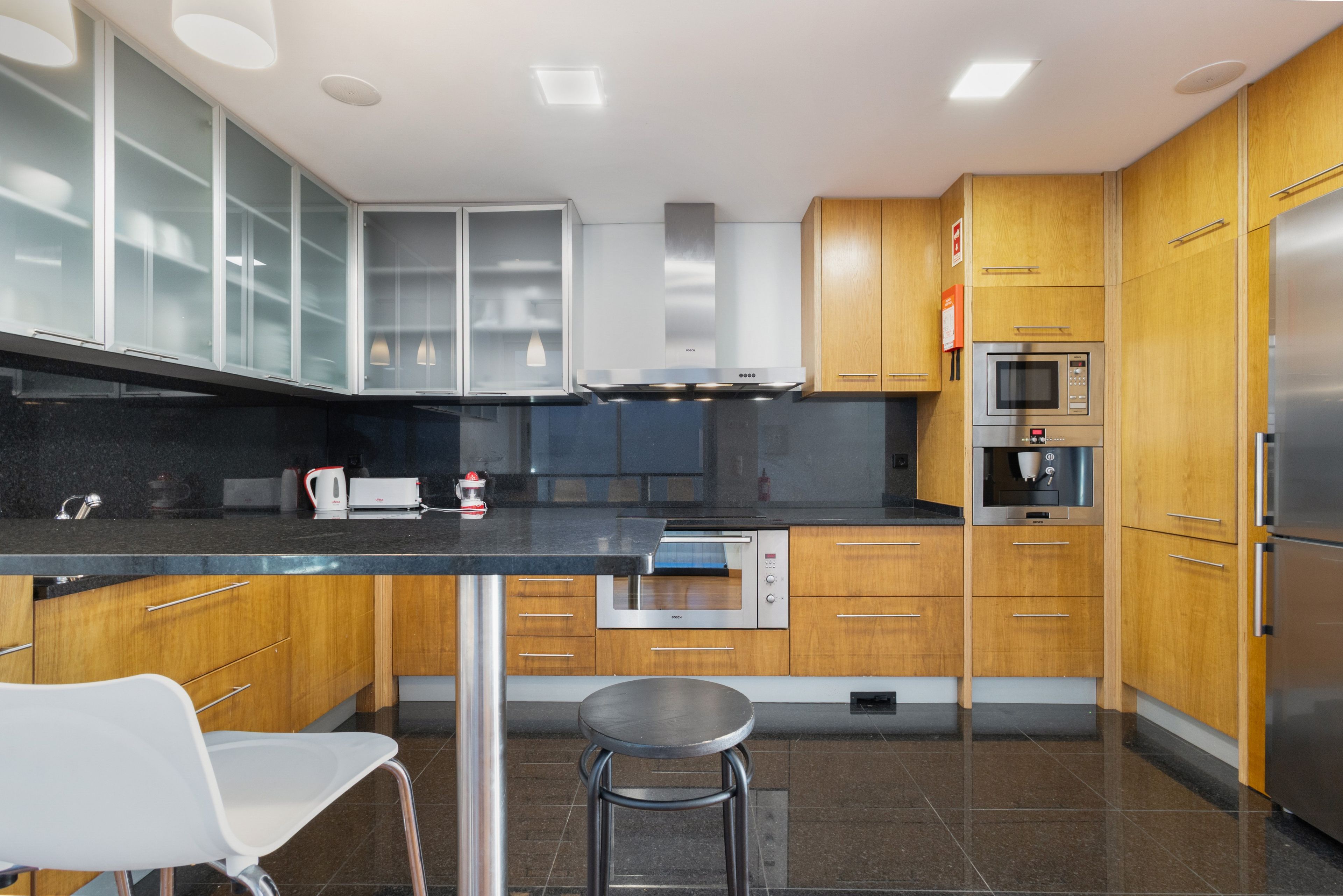 A modern kitchen with wooden cabinets, a stainless steel refrigerator, a built-in oven, and a coffee machine.