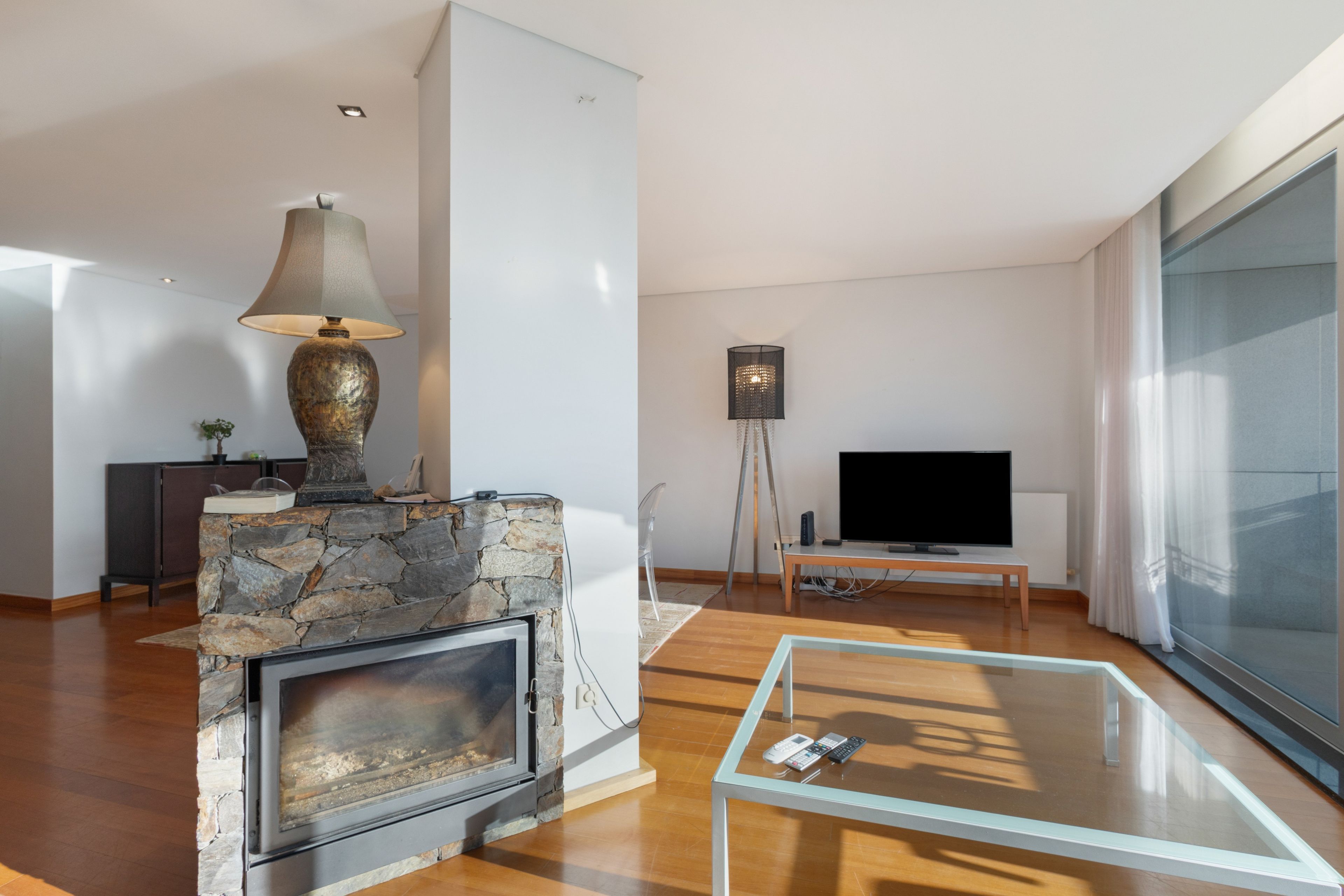 A modern living room with a stone fireplace, a glass coffee table, a television, and a floor lamp.