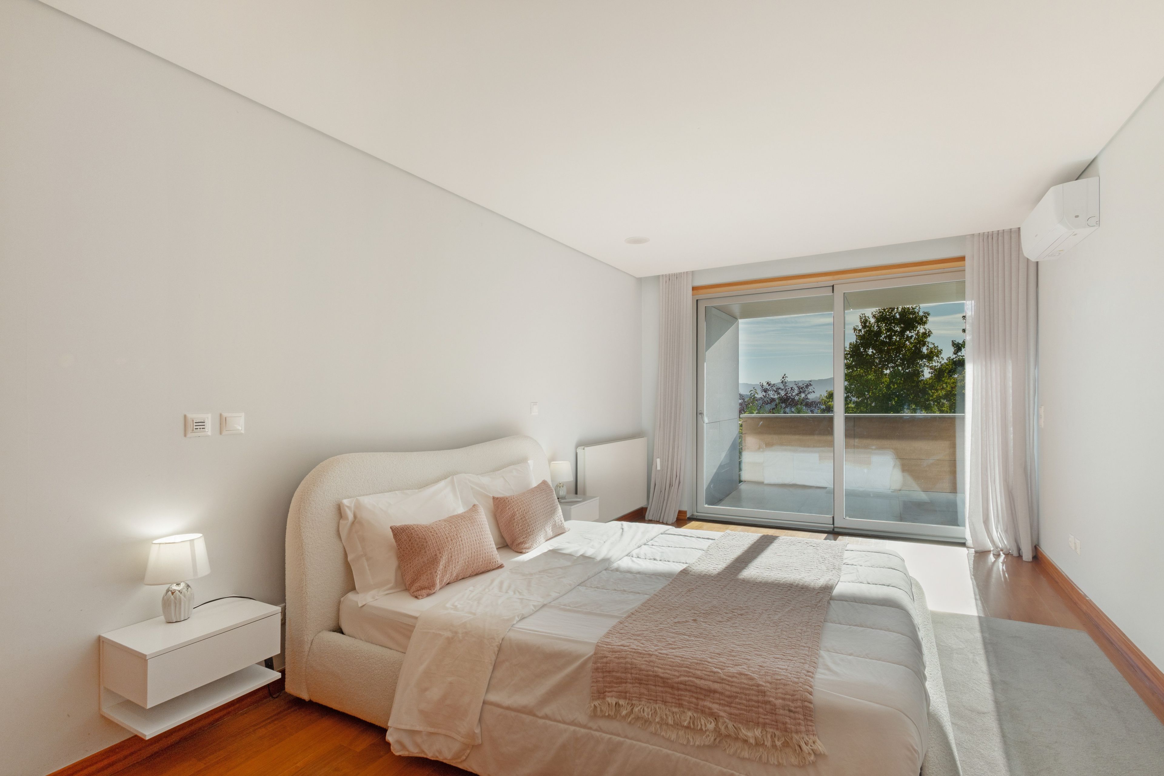 A modern bedroom with a large bed, a nightstand, a lamp, and a view of the outdoors through a sliding glass door.