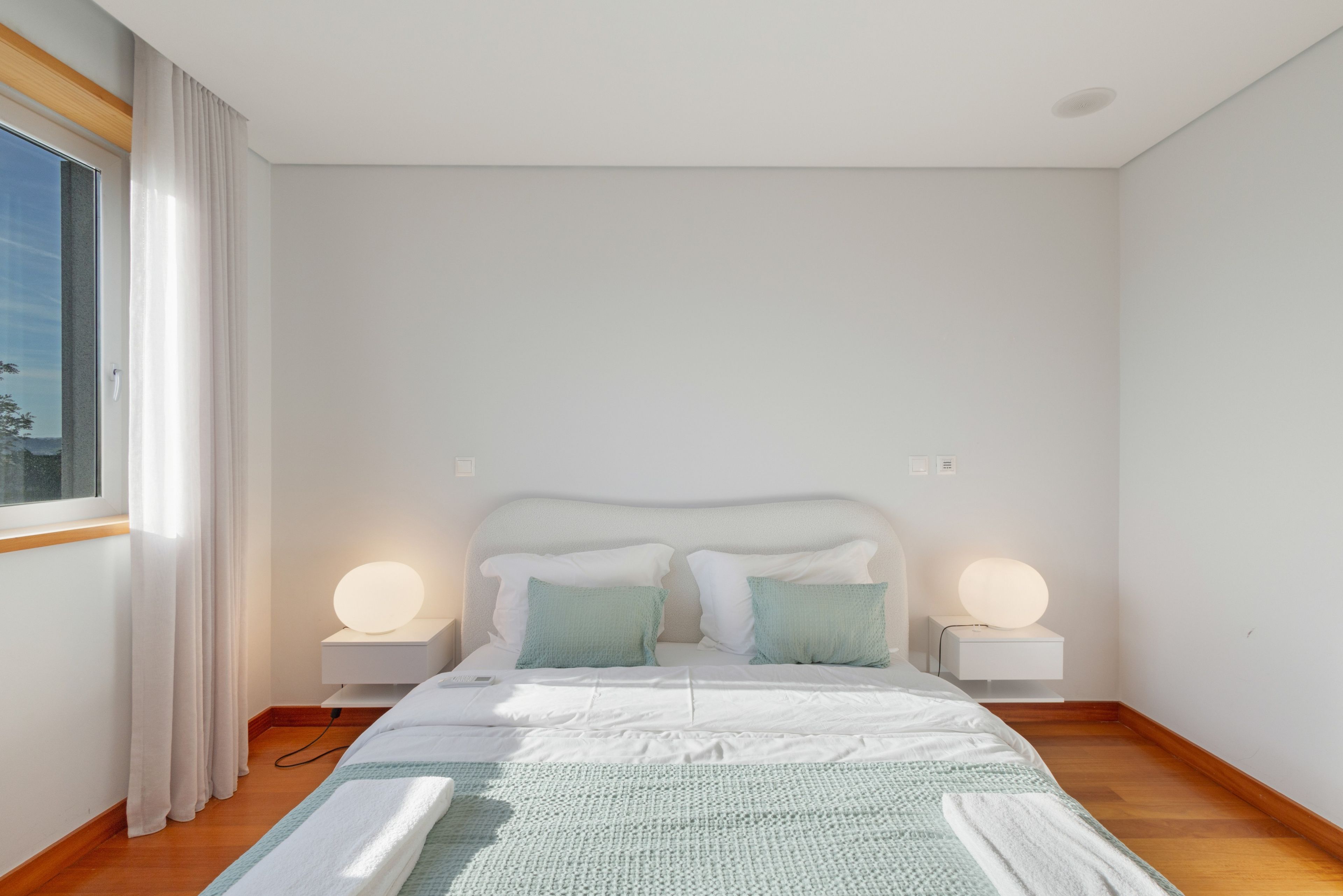 A minimalist bedroom with a large bed, two bedside lamps, and a view of the outdoors through a window.