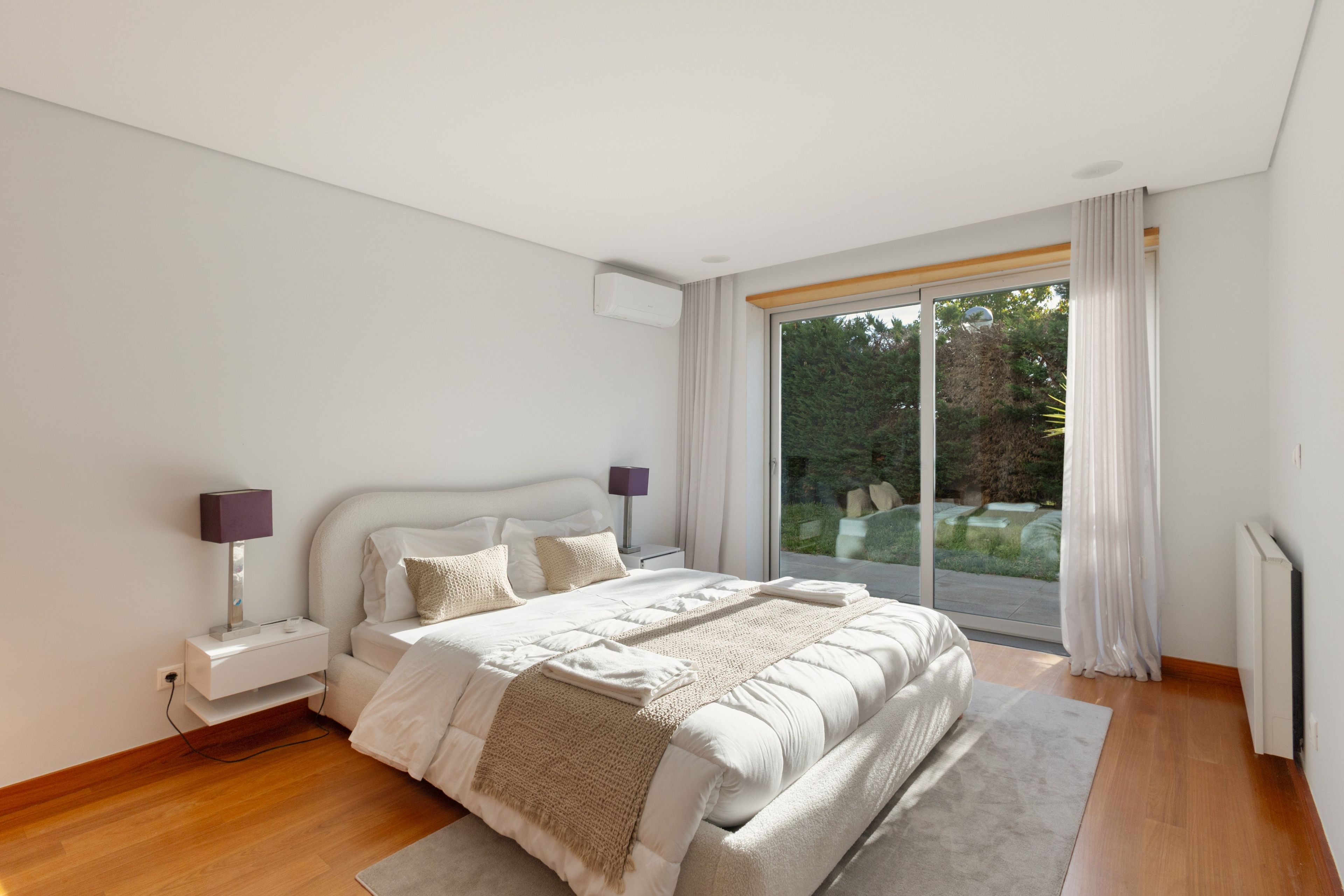 Una camera da letto moderna con un letto grande, lampade sui comodini e vista su un giardino tramite una porta scorrevole in vetro.