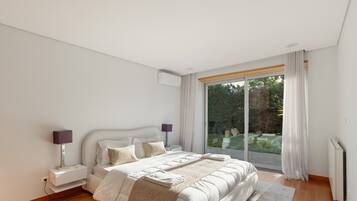 Ein modernes Schlafzimmer mit einem großen Bett, Nachttischlampen und Blick auf einen Garten durch eine Schiebetür.