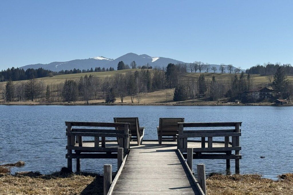 A wooden dock leading to a calm lake with benches, surrounded by hills and a clear sky.