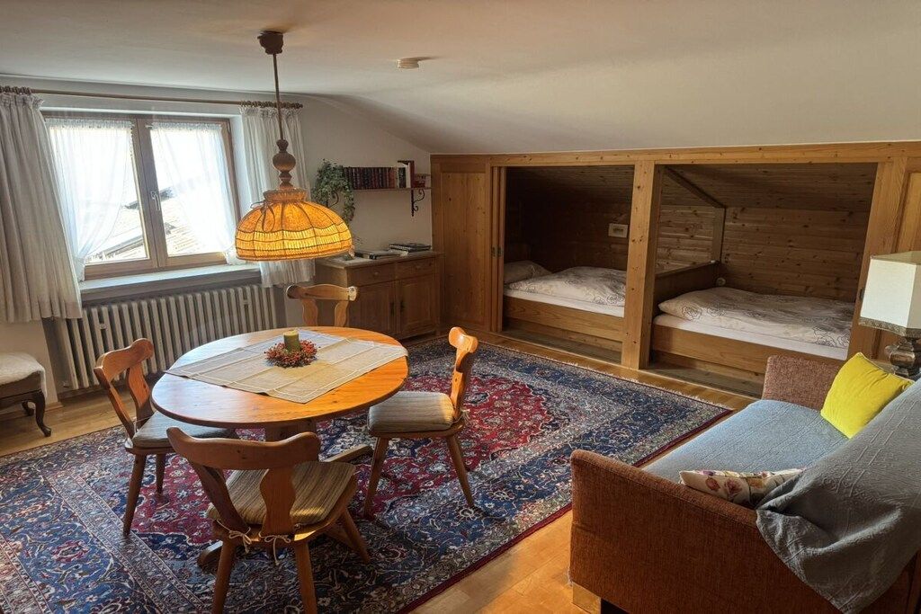 Een gezellige kamer met een houten hoogslaper, een eettafel met stoelen, een kamerkleed met dessin en een raam met gordijnen.