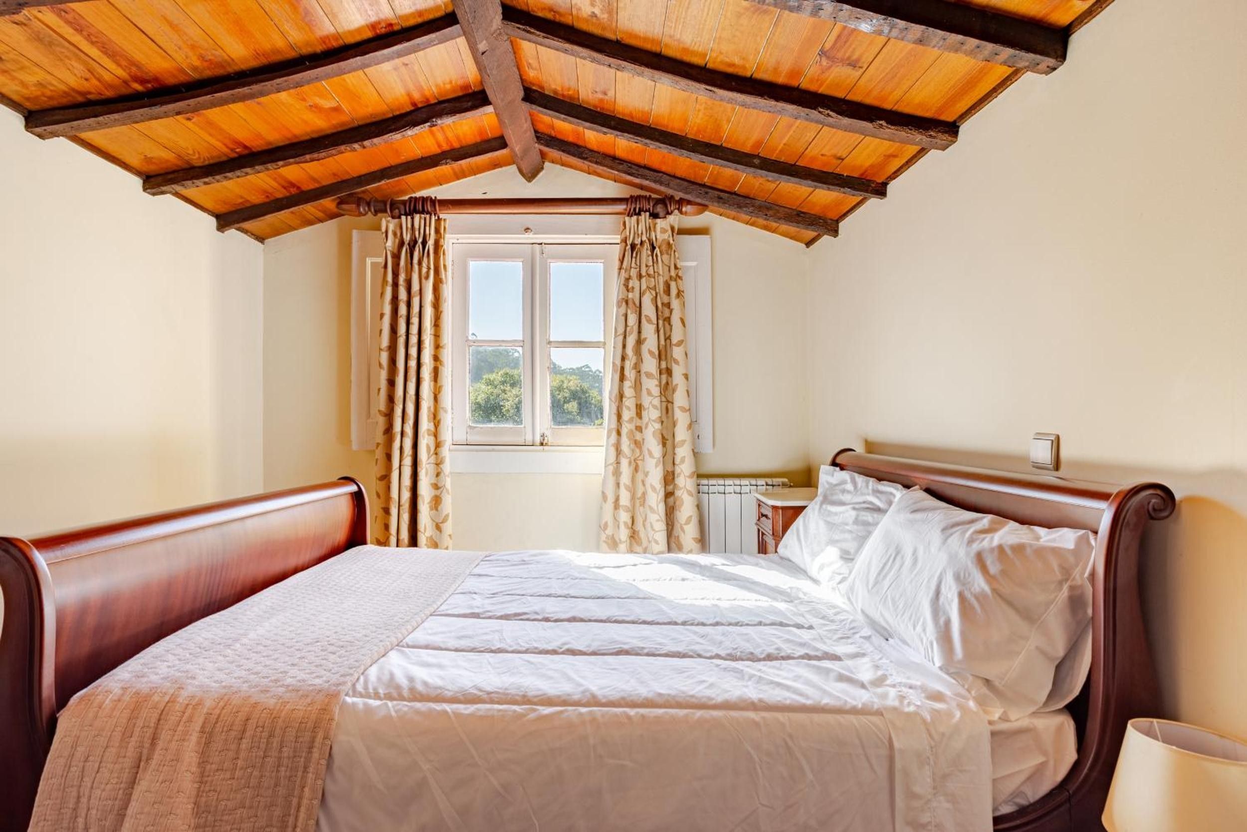 Ein Schlafzimmer mit Holzbalkendecke, einem Bett mit weißen Leinen, einem Fenster mit Vorhängen und einem Nachttisch.