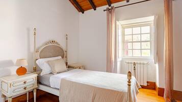 A bedroom with a wooden ceiling, a bed with a white headboard, a bedside table with a lamp, and a window with curtains.