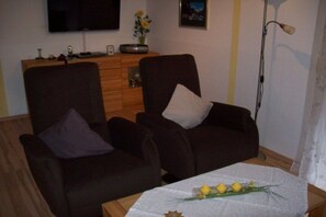 Ein Wohnzimmer mit zwei dunklen Sofas, einem kleinen Tisch mit dekorativer Dekoration, einem flachbildfernseher an der Wand und einer Holzkommode mit diversen Gegenständen.
