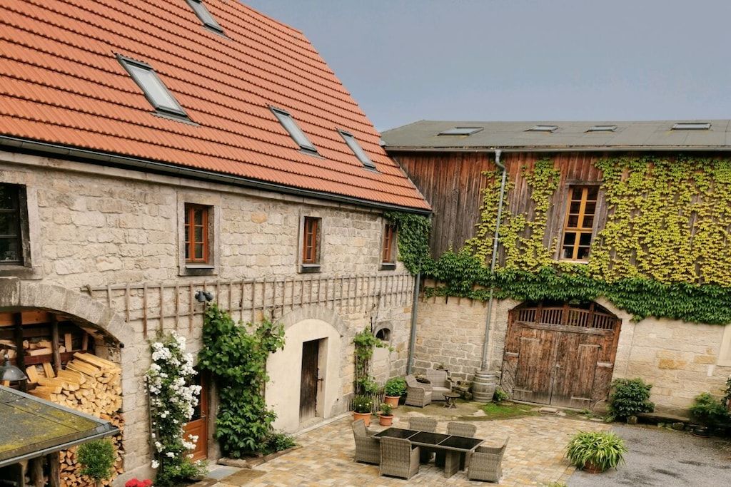 A stone building with a tiled roof, a courtyard with outdoor furniture, and a wooden door partially covered by ivy.