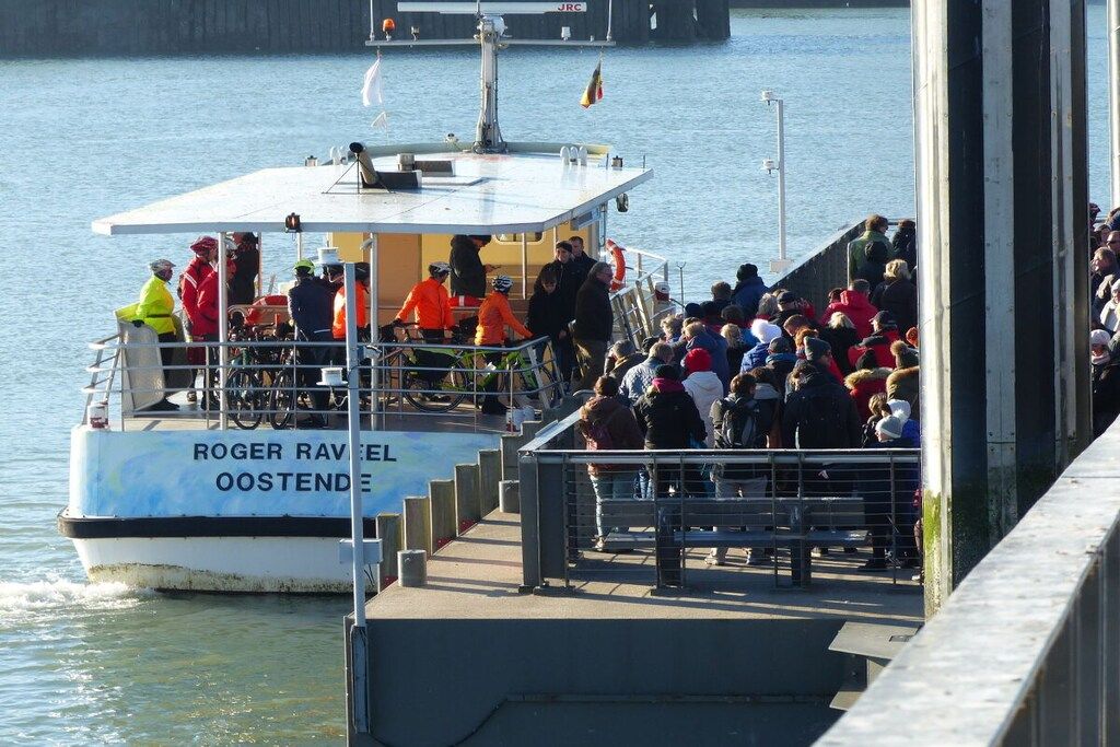 Ein Boot namens 'ROGER RAVEL' liegt an einem Steg fest, während sich Personen an Bord befinden und eine Menschenmenge vom Ufer aus zusieht.