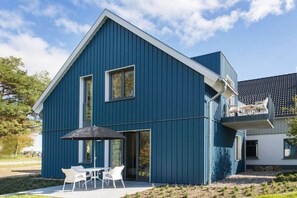 A modern two-story building with a blue exterior, a balcony with outdoor seating, and a patio area with a table and chairs. - House on the dike, harbor 3a H8 ground floor 48m² (Ostseebad Prerow)